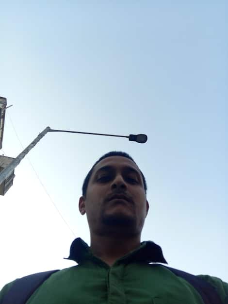 A close-up shot of a Peruvian engineer inspecting a wind turbine nacelle high above the ground. The engineer is wearing safety gear and using a tablet to monitor performance data. The background shows a vast wind farm with numerous turbines rotating, indicating technological expertise and renewable energy infrastructure development in Peru.