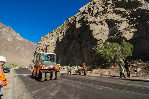 A construction site in the Peruvian Andes, showing heavy machinery working on a road or railway project. The mountain range in the background emphasizes the challenging terrain and the scale of the undertaking.
